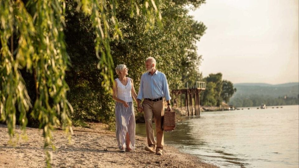 Mature couple strolling beside a river with a picnic basket