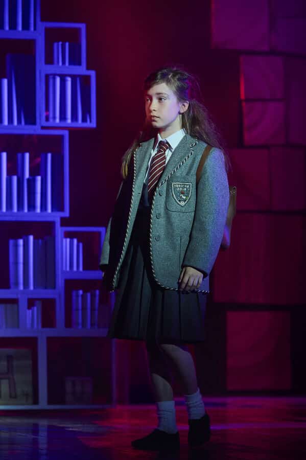 A young performer in school uniform beside a bookcase on stage