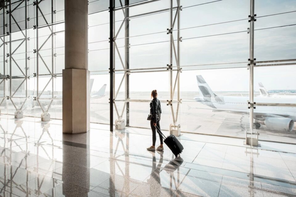 Woman with suitcase looking toward aircraft through terminal windows