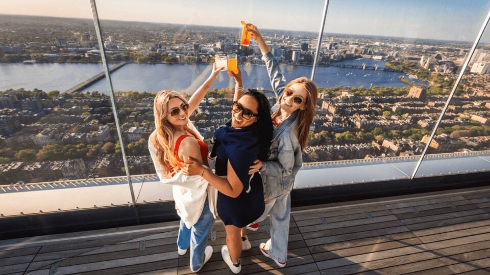 Friends raising drinks beside the glass observation deck with Boston and the Charles River behind them